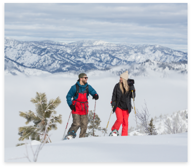 Two people snowshoeing on a snowy mountain trail with snow-covered trees in the foreground and expansive mountain ranges in the background under a cloudy sky.