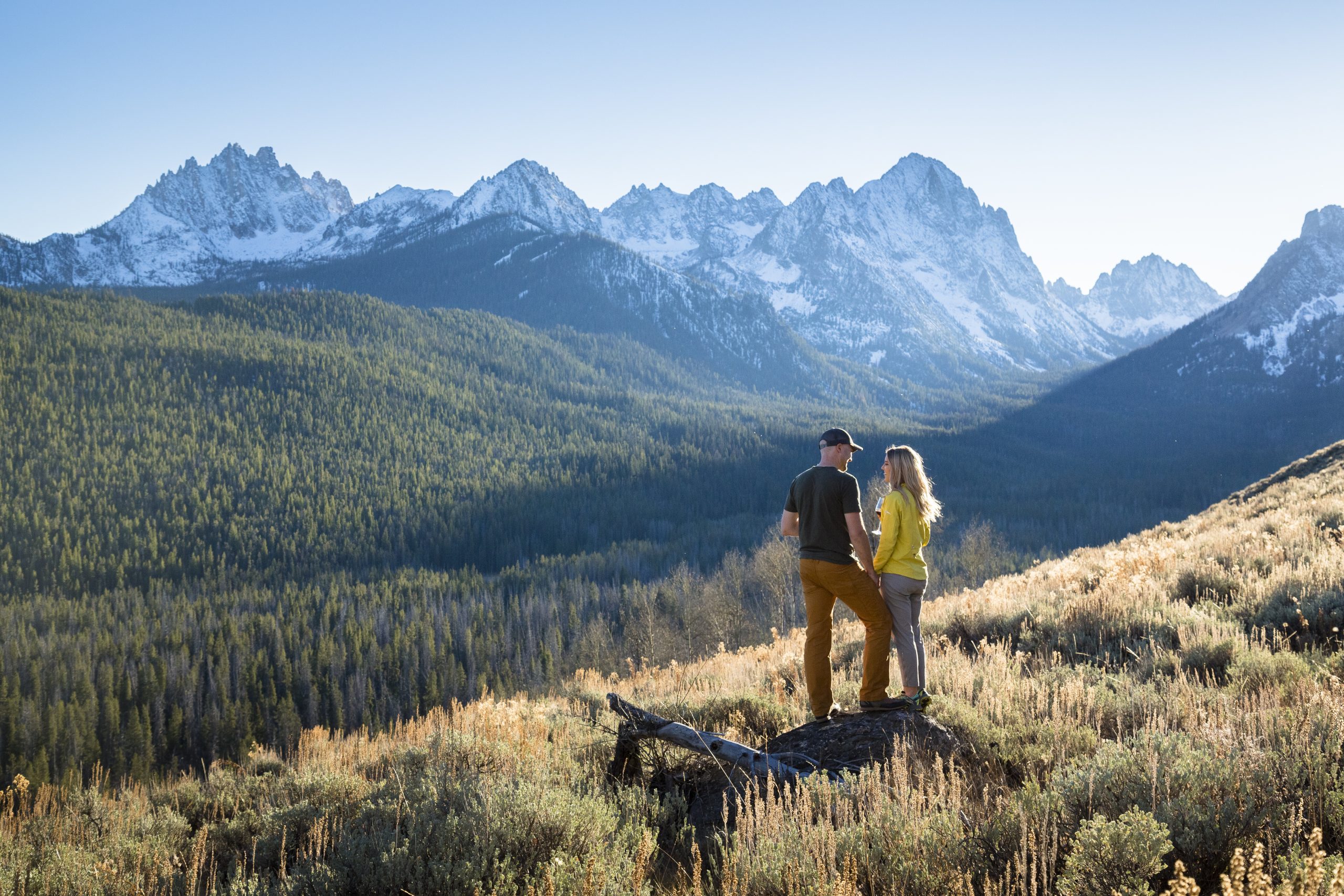 Two people standing on a hill covered in tall grass overlooking a vast landscape of tall grass and snow-covered mountains in the distance.