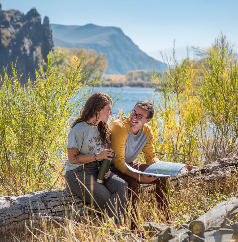 Two people reading a map while sitting on a fallen log amidst some tall bushes.