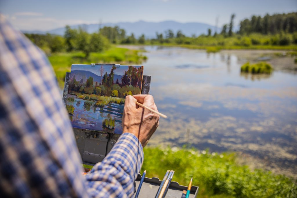 A close-up view of a person painting the scenery before them—a body of water surrounded by a landscape of tall grass and trees.