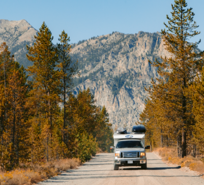 An RV en route to Alice Lake, navigating a road lined with trees with fall foliage and a series of mountains in the background.