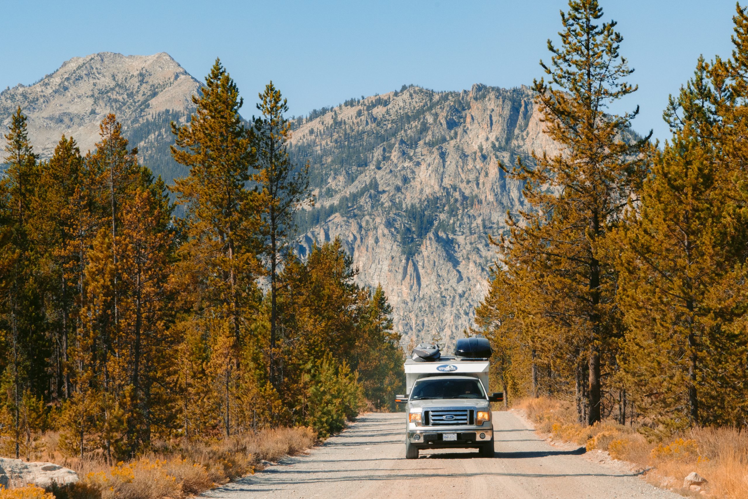 An RV en route to Alice Lake, navigating a road lined with trees with fall foliage and a series of mountains in the background.