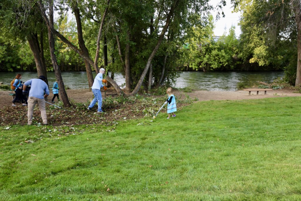 A group of people raking tree branches and picking up trash in a park along a river.